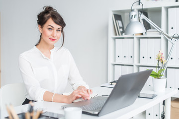 Wall Mural - Close-up portrait of a businesswoman at her workplace working with pc, looking in camera, wearing office suit.