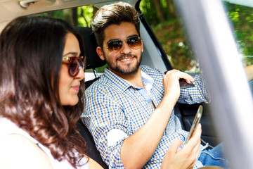 Wall Mural - Portrait of young couple driving a car. They are looking at mobile phone. 