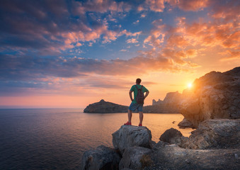 Wall Mural - Young standing man with backpack on the stone on the seashore at colorful sunset sky. Beautiful landscape with sporty man, rocks, sea and clouds at sunset. Sport, lifestyle background. Travel.