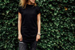 Cropped image of young hipster girl with curly hair wearing a blank black t-shirt and black jeans. mock-up of blank t-shirt. plants background. empty place for your text o design.