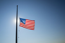 American Flag Flies At Half Mast Backlit By The Sun In Bright Blue Sky