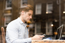 Window View Of Young Businessman Reading Smartphone Texts In Cafe
