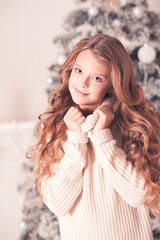 Smiling teenage girl with long blonde curly hair wearing cozy knitted sweater over Christmas tree in room. Looking at camera.