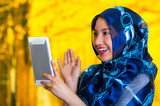 Fototapeta Panele - Beautiful young muslim woman wearing blue colored hijab, holding up tablet staring at screen, autumn forest background