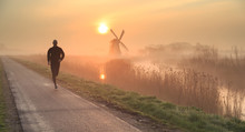 Man Running In The Foggy, Dutch Countryside Near A Windmill During A Tranquil Sunrise. Shallow D.O.F.