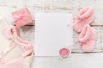 Baby girl clothes, blank card with wax seal and small present box on white wooden background. Flat lay. Owerhad view