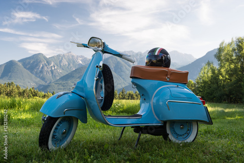 Cuadros en Lienzo Blaue Vespa vor Bergkulisse auf Feld