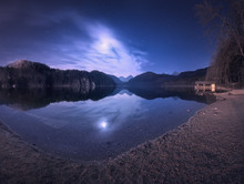 Colorful Night Landscape With Lake, Mountains, Forest, Stars, Full Moon, Purple Sky And Clouds Reflected In Water. Spring Night In Alpsee Lake In Germany. Panoramic Photo. Nature Background