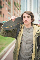 Wall Mural - Half length of a young handsome caucasian man with mustache posing with his skateboard in the street of the city looking right - freshness, youth, carefreeness concept