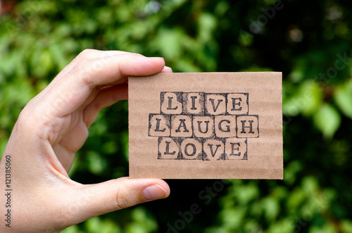 Woman hand holding cardboard card with words Live Laugh Love made by black alphabet stamps opposite green floral background Poster