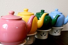 Teapots / Pink, Yellow, Green And Blue Teapots With Warmers, Lined Up In A Row.