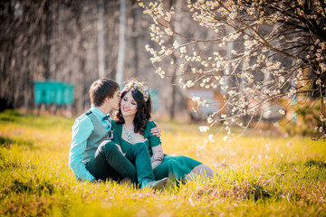 Young beautiful couple kissing and hugging near trees with blossom in summer park near stump on the grass. Woman in green dress and wreath. Man in blue shirt and gray waistcoat