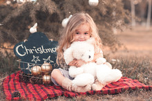 Smiling Baby Girl 4-5 Year Old Holding Teddy Bear Toy Sitting With Christmas Decorations Outdoors. Looking At Camera.