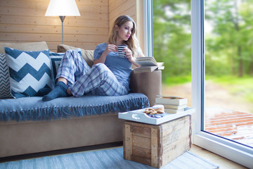 Young beautiful blonde woman with cup of coffee and book sitting home by the window. Summer spring green woods view. Lazy day off concept