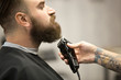 © fizkes - Close-up side view portrait of handsome young bearded caucasian man getting beard grooming in modern barbershop. Hairdresser with tattoo 'one love' serving client, making beard haircut using shaver