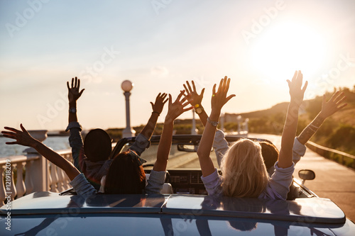 Fotografie, Obraz Back view of happy friends driving cabriolet with raised hands