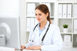 © rogerphoto - Young brunette female doctor sitting at the table and working at hospital office.  Health care, insurance and help concept. Physician ready to examine patient