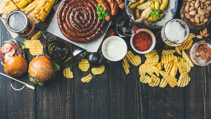 Wall Mural - Beer and snack set. Octoberfest food frame concept. Variety of beers, grilled sausages, burgers, corn, fried potatoes, chips, salted almonds and sauces on dark wooden background. Top view, copy space