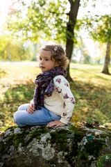 Portrait of a beautiful little girl playing in the autumn park. Thoughtful child. Cute kid sitting on a big stone with moss. Sunny weather, fresh air, trees with yellow leaves. Healthy lifestyle.