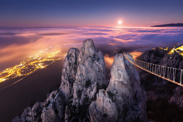 Wall Mural - Mountain landscape with rising full moon and fog in the city at night. Beautiful night landscape with mountain peak, bridge, low clouds, moon and city lights. High rocks against sky with purple clouds