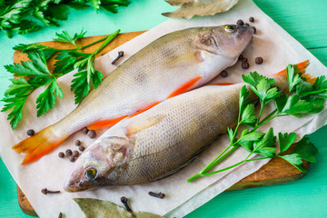 Two raw perch on rustic cutting board with fresh herbs and spices on green wooden table