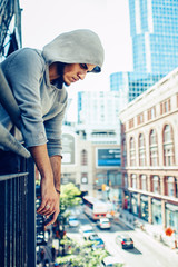 Wall Mural - Conceptual art portrait of beautiful handsome pensive sad tired gothic young middle east brunette man with beard in hoodie, standing on staircase in city urban looking down on street