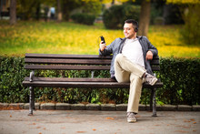 Portrait Of A Relaxed Young Man Sitting On Bench In Park And Listening To Music On Headphone