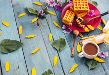 Blue Wood Background, Dotted With Yellow Flower Petals And Green Leaves. Peeled Banana. A Cup Of Coffee, Maroon Plate Of Biscuits For Breakfast.