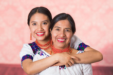 Beautiful Hispanic Mother And Daughter Wearing Traditional Andean Clothing, Embracing While Posing Happily Together Interacting For Camera, Pink Studio Background