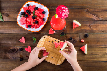 Wall Mural - Plate with salad of watermelon, blackberries, mint. hand cut hearts of watermelon on a wooden board. Watermelon fresh in a glass with an umbrella. On a wooden background.