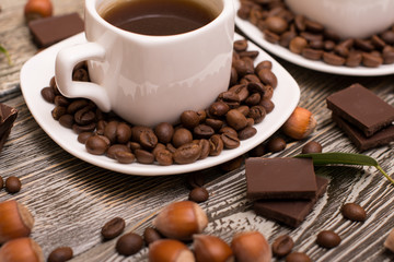 two small white cups of coffee with cocoa beans, slices of chocolate, hazelnuts and green leaves on wooden background