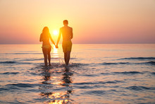 Silhouette Of Two Lovers Walking Hand To Hand On The Beach At Sunset Time