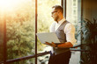 © SFIO CRACHO - Stylish Bearded Businessman Wearing Glasses White Shirt Waistcoat Holding Modern Laptop Hands Looking Panoramic Window.Man Working Office Startup Project Workplace.Sunset Filter Effect.Blurred.