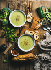 Two bowls of homemade pea, broccoli and zucchini cream soup served with fresh baguette and vegetables on wooden board over rustic background, top view, vertical composition