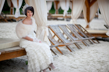 Wall Mural - Thoughtful bride sits enveloped in white fur on the beach