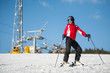 © anatoliy_gleb - Woman wearing helmet, red jacket and ski goggles standing with skis on mountain top at a winter resort in sunny day with ski lifts and blue sky in background.