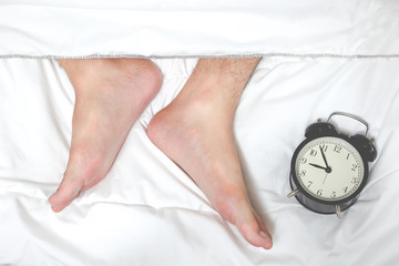 feet of lazy man on white bed & alarm clock. wake up late