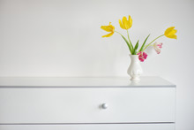 Interior Design Scene With A White Modern Commode And A Vase Of Yellow And Pink Tulips On A Pale Grey Wall
