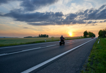 Motion blur motorcycle riding on asphalt road towards the horizon in rural landscape at sunset. Red car in the distance.