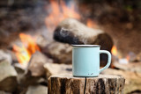 Fototapeta  - Blue enamel cup of hot steaming coffee sitting on an old log by an outdoor campfire. Extreme shallow depth of field with selective focus on mug.