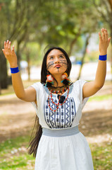 Wall Mural - Beautiful Amazonian woman with indigenous facial paint and white traditional dress posing happily for camera, interacting holding out arms in park environment, forest background