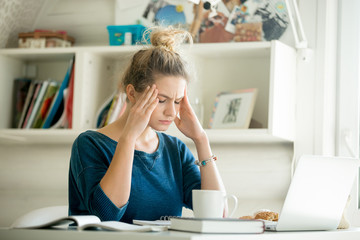 Portrait of an attractive woman at table with cup and laptop, book, notebook on it, hands at her temples. Bookshelf at the background. Concept photo