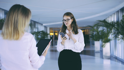 Wall Mural - Two business women standing in the lobby and checking email on the gadgets.Brunette in glasses and a white blouse holding a smartphone,in the foreground a girl with the tablet computer.