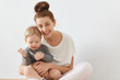 © WHstudio Leushin N - Beautiful portrait of young mother and child sitting together on white background. Happy Caucasian female with bunch of brown hair in white clothes holding baby in her arms, sincerely smiling.