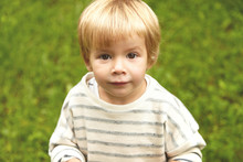 Charming Close-up Innocent Portrait Of Little Innocent Kid. Calm Caucasian Boy With Blond Hair Round Brown Eyes Looking At Camera. Five-years Old Infant Standing Still On Fresh Green Grass Background.