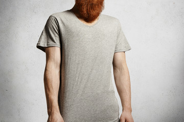 Trendy redhead student wearing blank copy space t-shirt spending free time indoors. Caucasian model with long red beard dressed casually posing in studio against blank concrete wall background