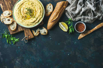 Homemade hummus dip in blue ceramic bowl with fresh baguette, lemon, herbs and spices over dark blue plywood background, top view, copy space