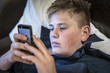 Young teenage boy reading a text message on his mobile phone as he relaxes on a pillow, close up view of his face