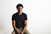 Friendly Smiling Attractive Young Man In Unlabeled Cotton Black T-shirt And Jeans Sitting On A Chair And Looking Into Camera Isolated On White