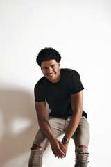 Wall Mural - Smiling athletic young African American man in blank black cotton t-shirt and jeans sitting at a white wall